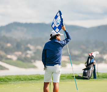 A young golfer in a navy jacket holds the flagstick on a green overlooking a hazy mountain landscape
