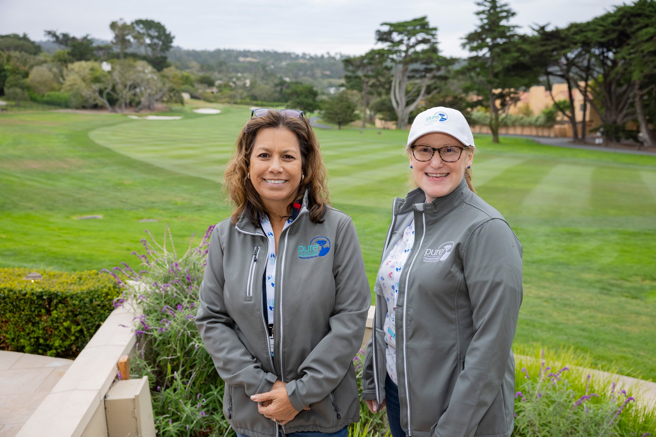 Two female tournament volunteers in grey jackets and caps smiling together on a lush green course