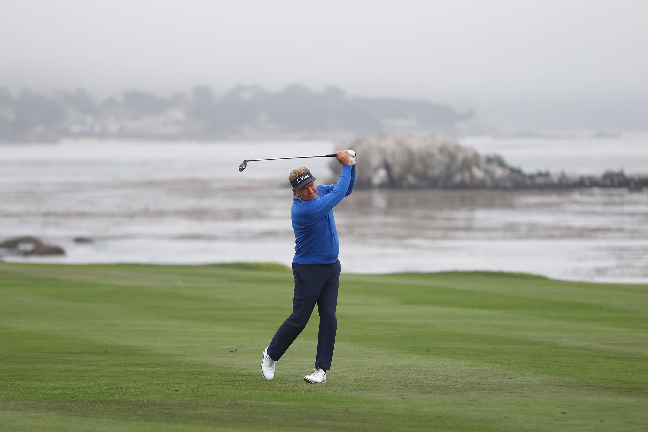Ken Duke finishing a golf swing on a vibrant green fairway with a rocky coastline in the misty background