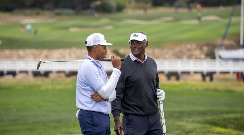 A young golfer and a senior professional in a grey sweater share a conversation on the fairway