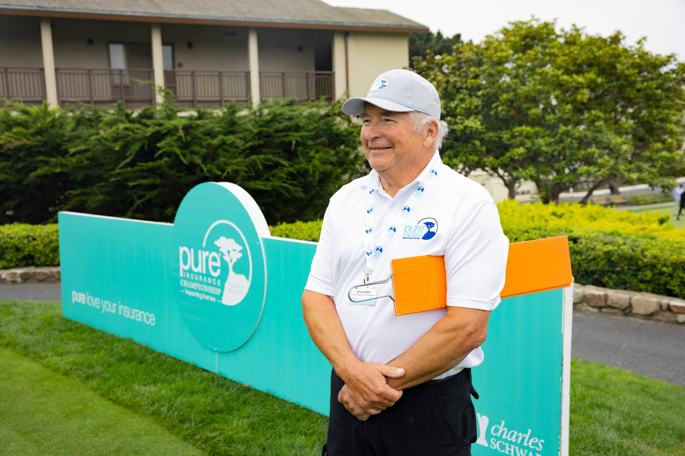 Smiling volunteer in a white PURE Insurance polo and grey cap holding an orange paddle near a tournament sign