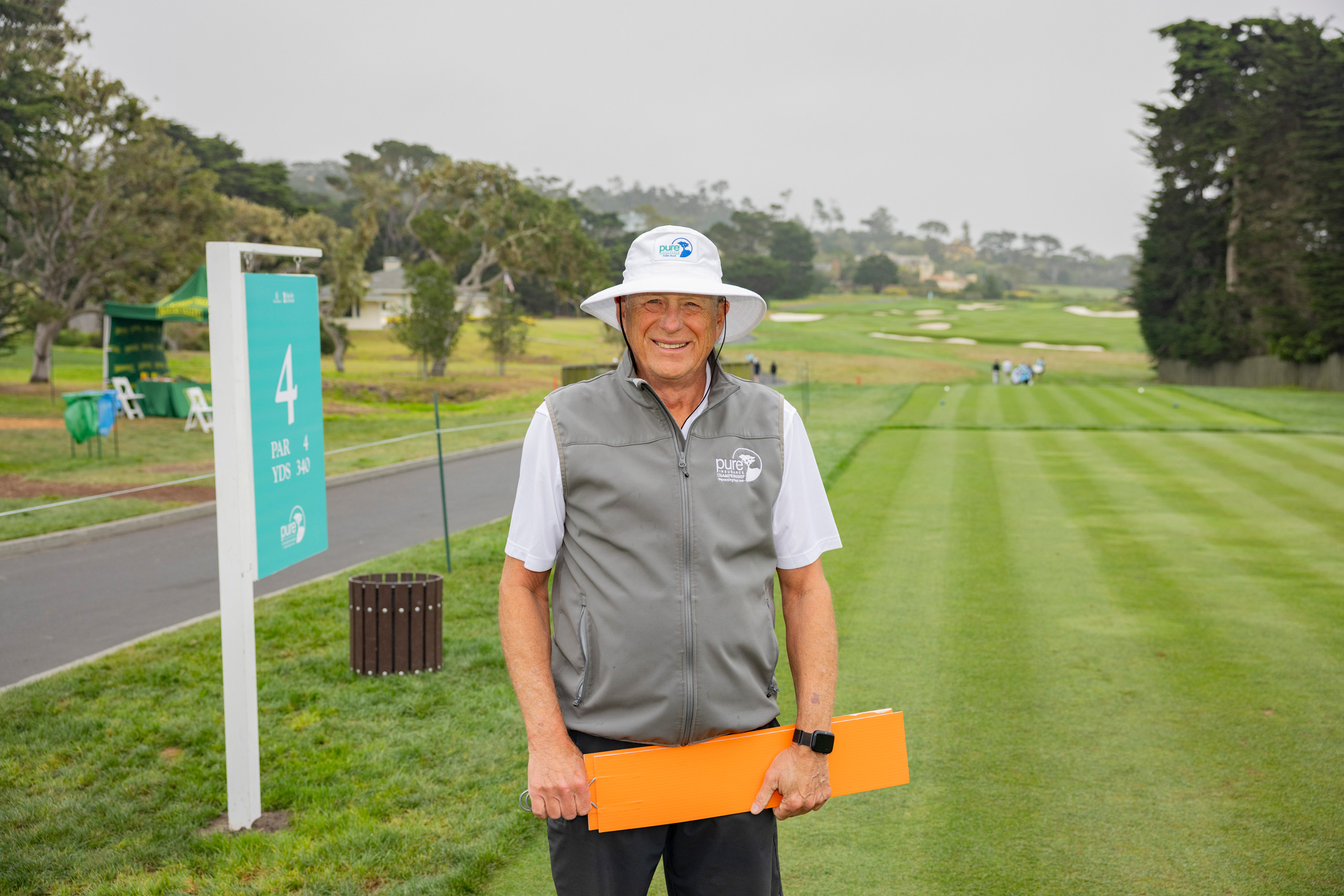 Volunteer in a white sun hat and grey vest holding an orange signal paddle by the Hole 4 yardage sign