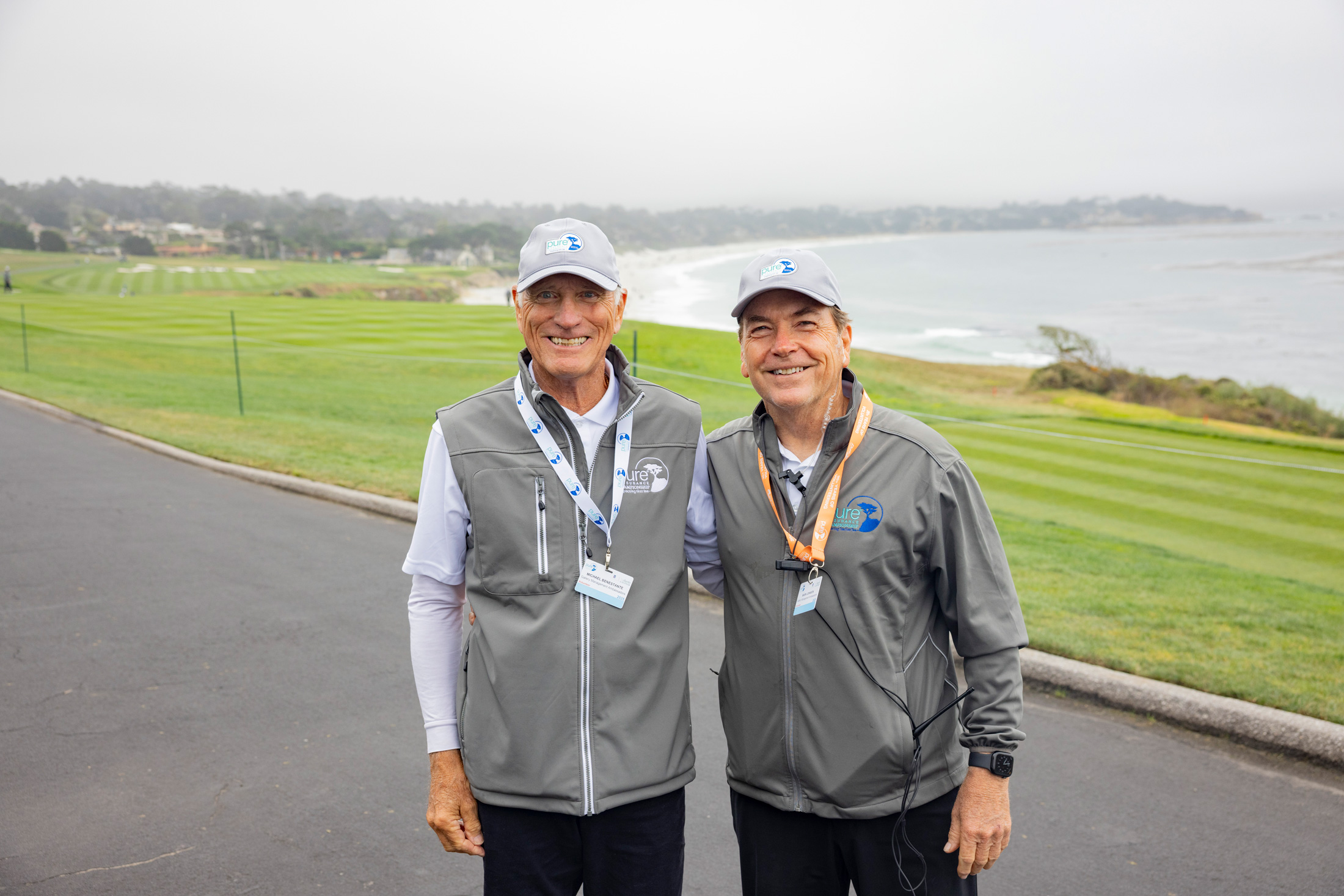 Two smiling male volunteers in grey tournament vests and caps posing together on a coastal path