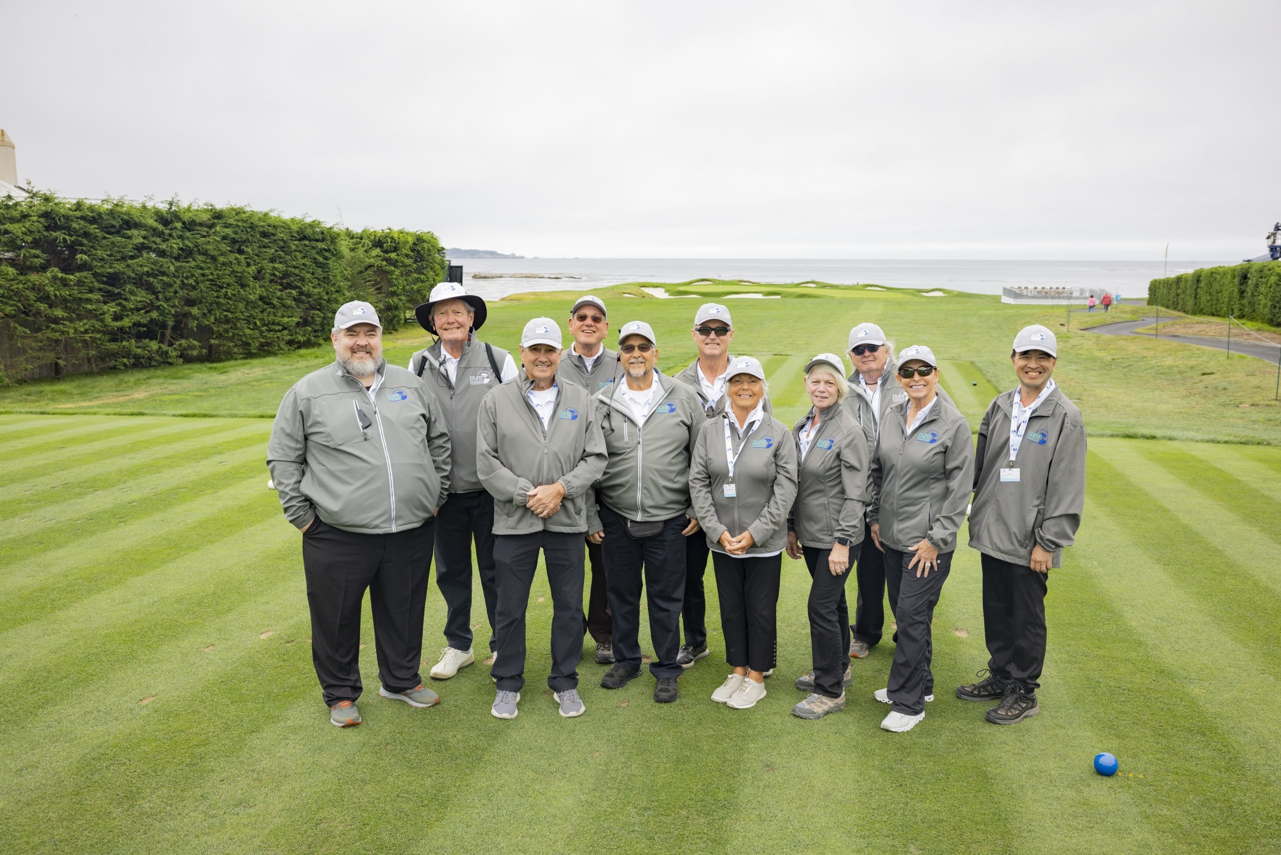 A large group of tournament volunteers in matching grey jackets and white caps posing on a green