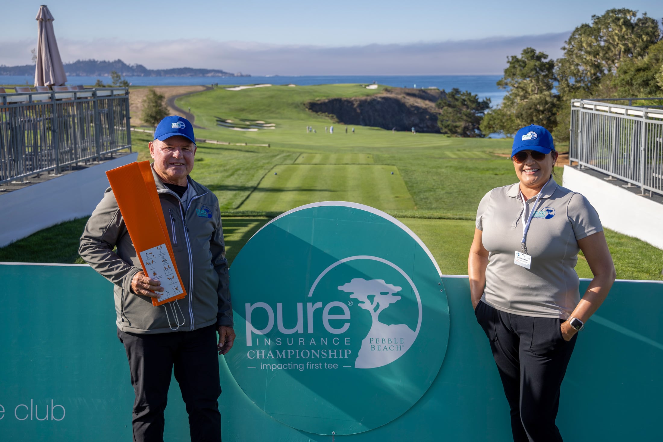 Two volunteers posing behind a large circular PURE Insurance Championship sign with Hole 6 cliffs in the background