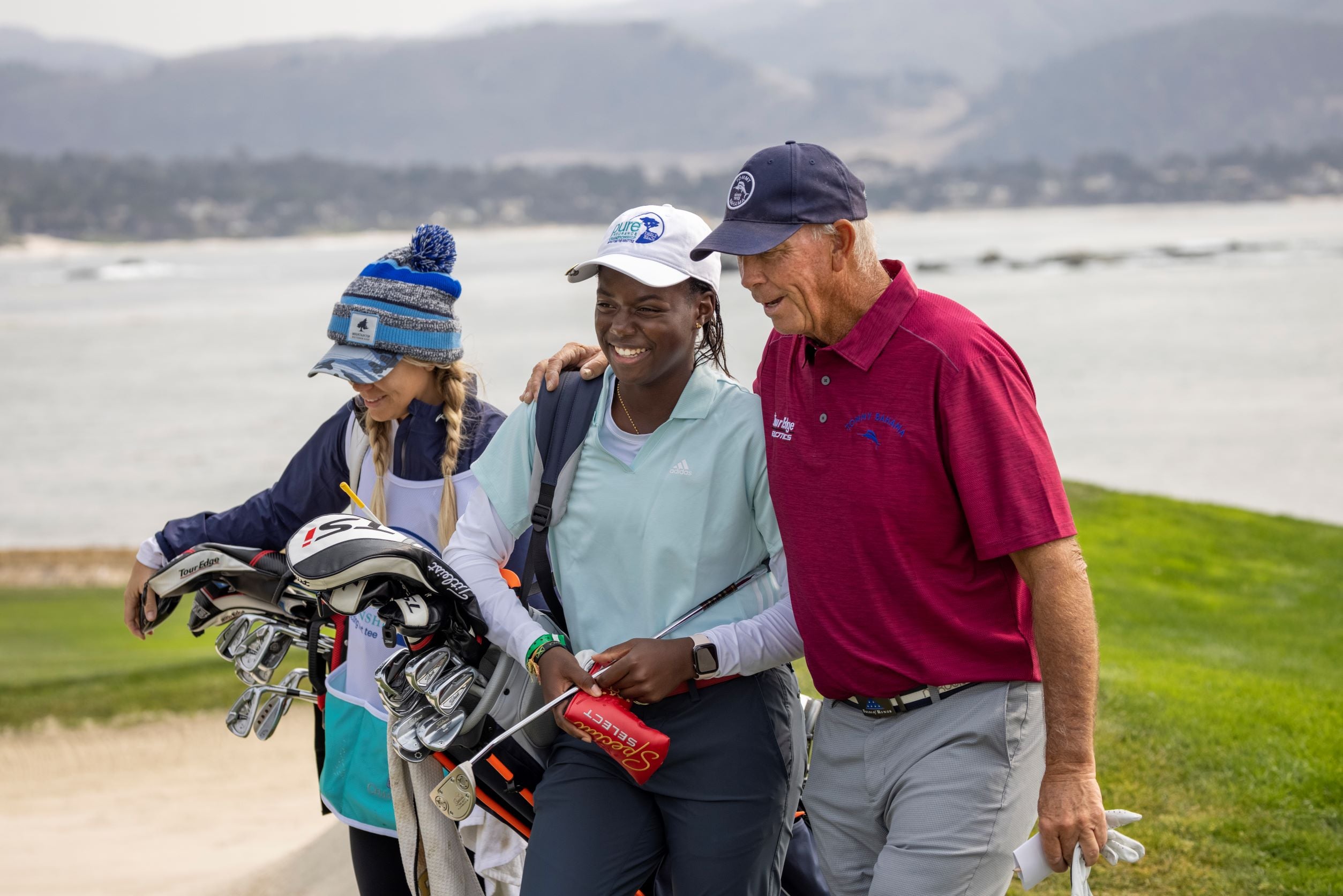 A smiling young golfer walks along the coast with her caddy and a senior professional mentor