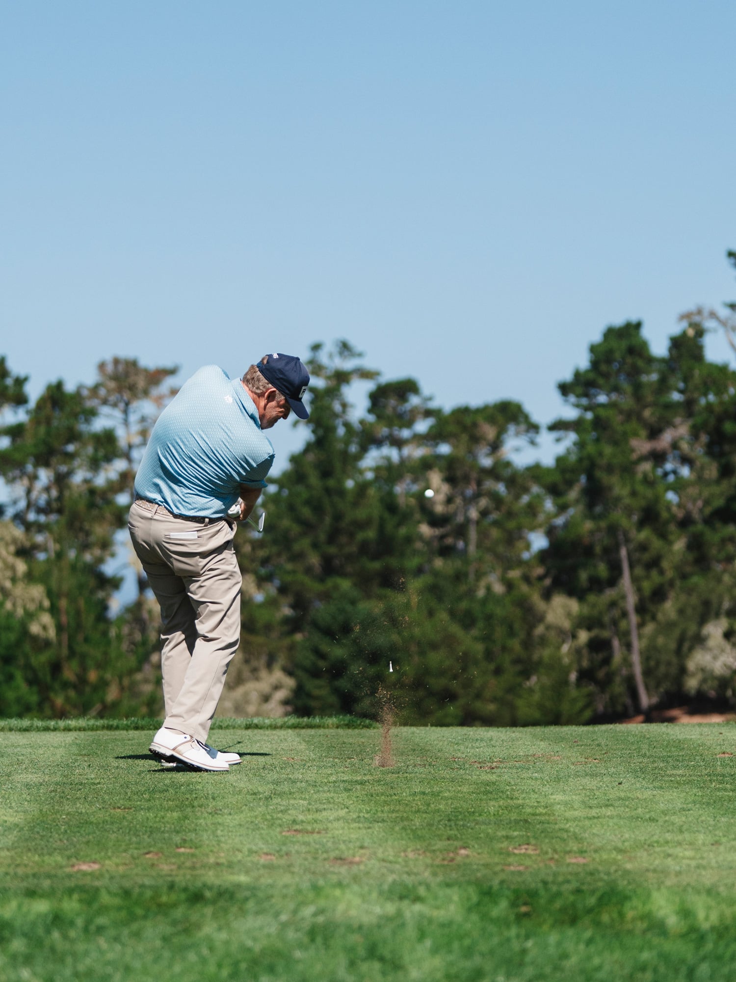 A professional golfer in a light blue shirt completing a full swing on a tree-lined tee box