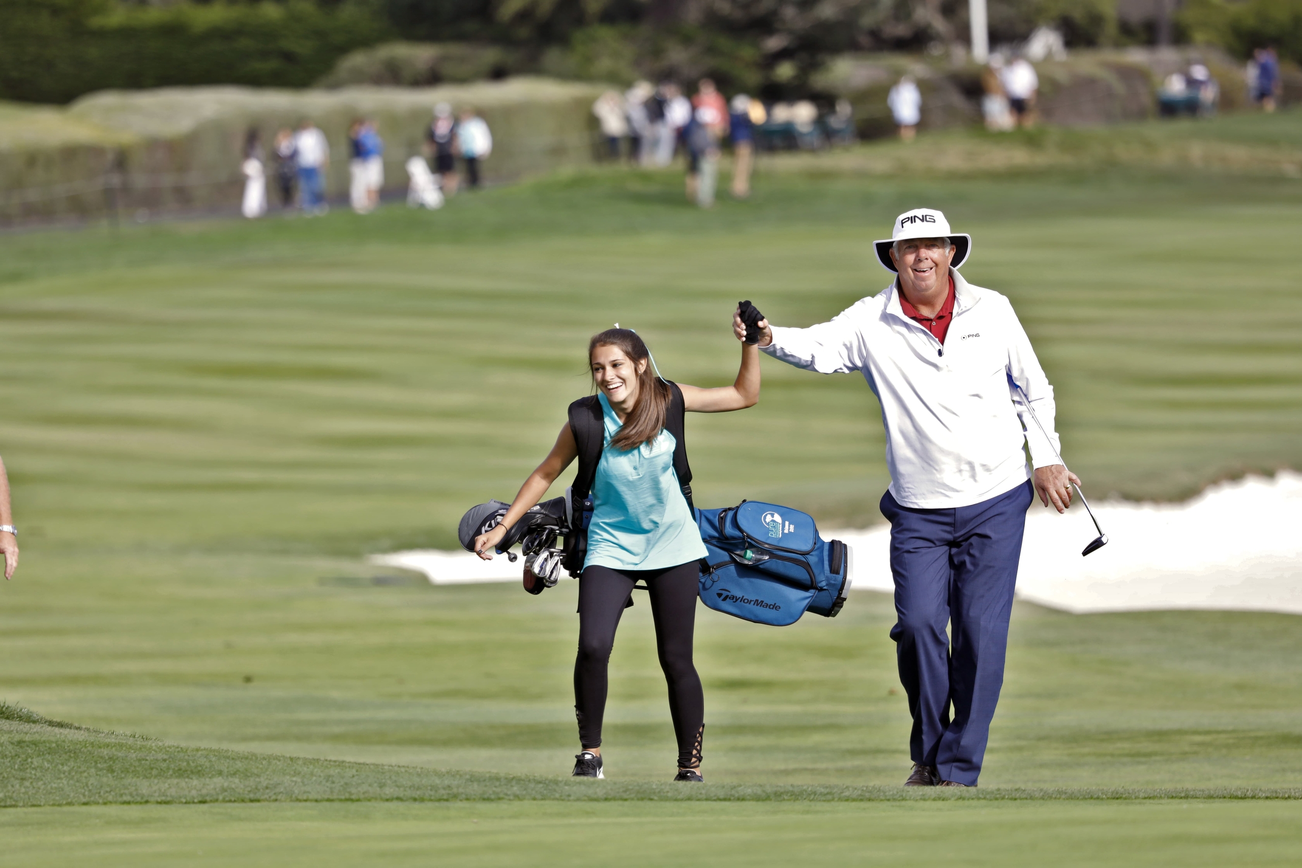A smiling professional golfer holds the hand of a young female participant carrying her golf bag