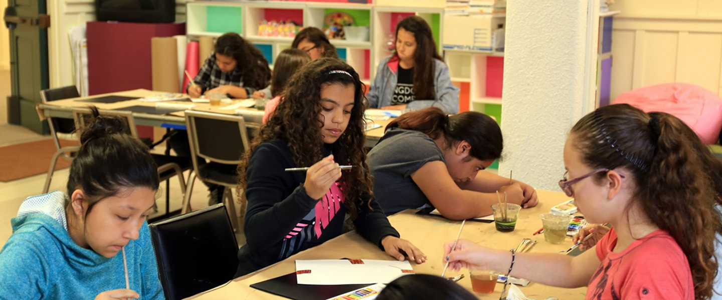 Diverse group of young students sitting at long tables focused on an indoor art or painting activity