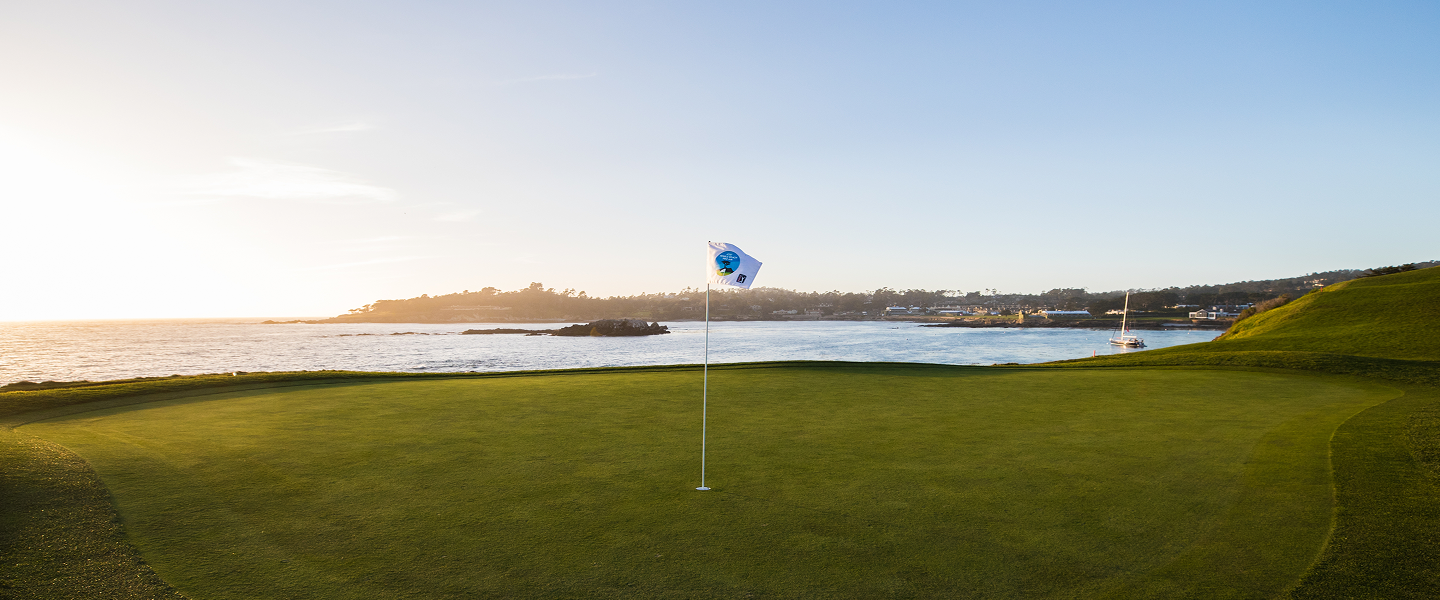 Wide-angle sunset view of a green at Pebble Beach with a tournament flag overlooking the bay