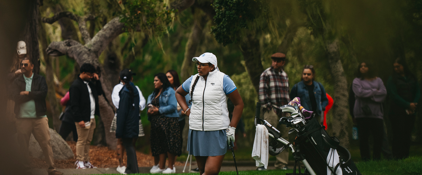 A female golfer in a white vest and blue skirt stands near her golf bag among a crowd of spectators