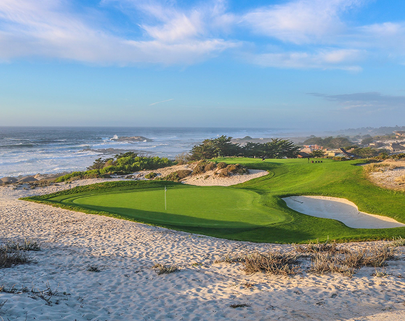 Green golf hole surrounded by white sand dunes and shrubs next to the crashing ocean waves