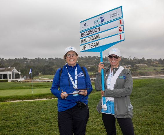 Two female volunteers; one takes notes while the other holds a scoring placard for Bransdon and teams