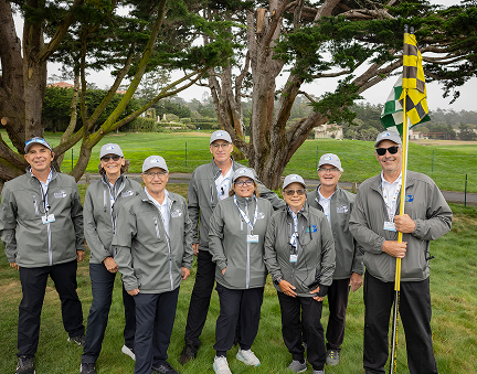 Group of eight volunteers in grey tournament gear posing in front of a large cypress tree on the course