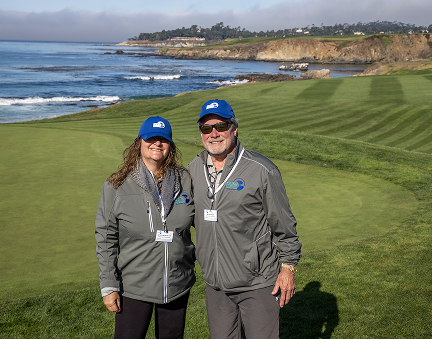 Two smiling volunteers in blue caps and grey jackets posing on a green with the blue ocean behind them