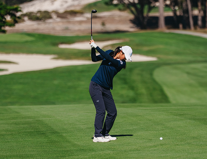 Female golfer in a navy long-sleeve shirt and white cap mid-swing on a bright green fairway