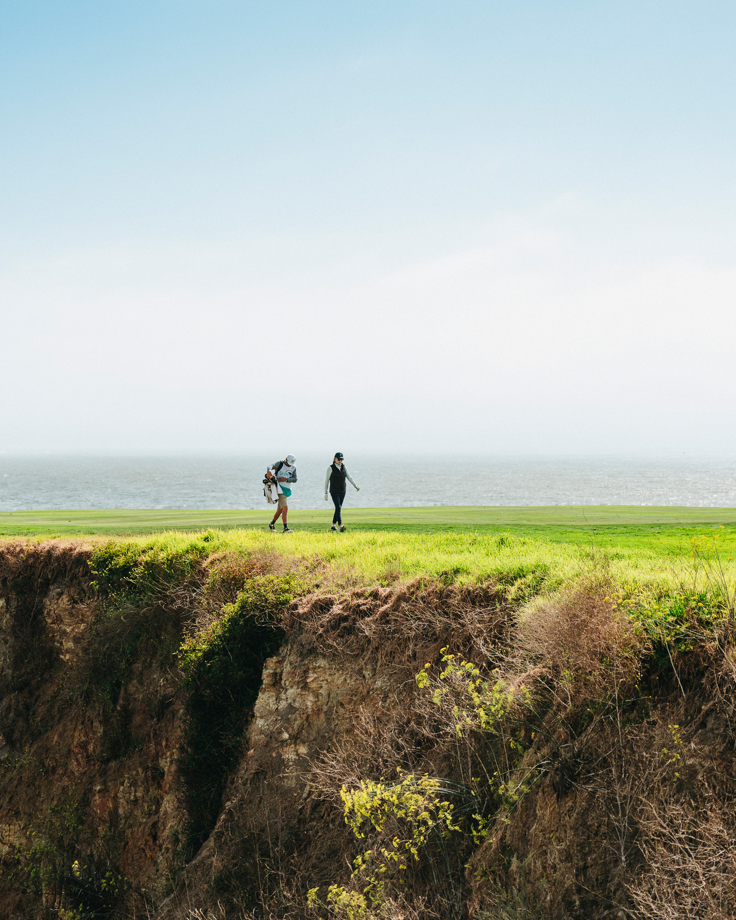 Two golfers walking across a green fairway along a high coastal cliff overlooking the ocean
