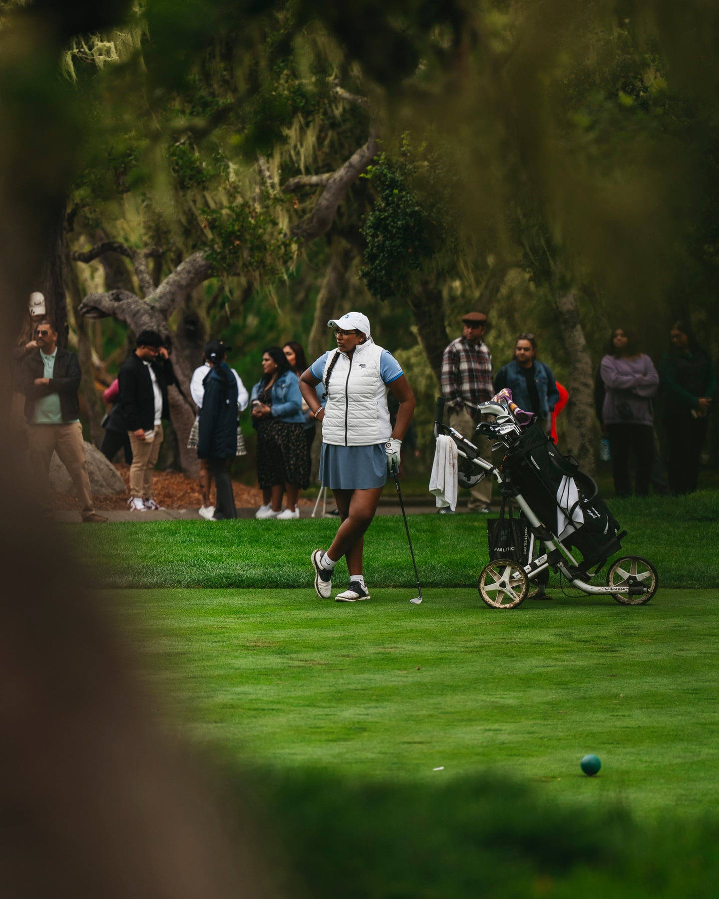 female golfer stands with her hands on her hips next to her bag on a tree-lined fairway