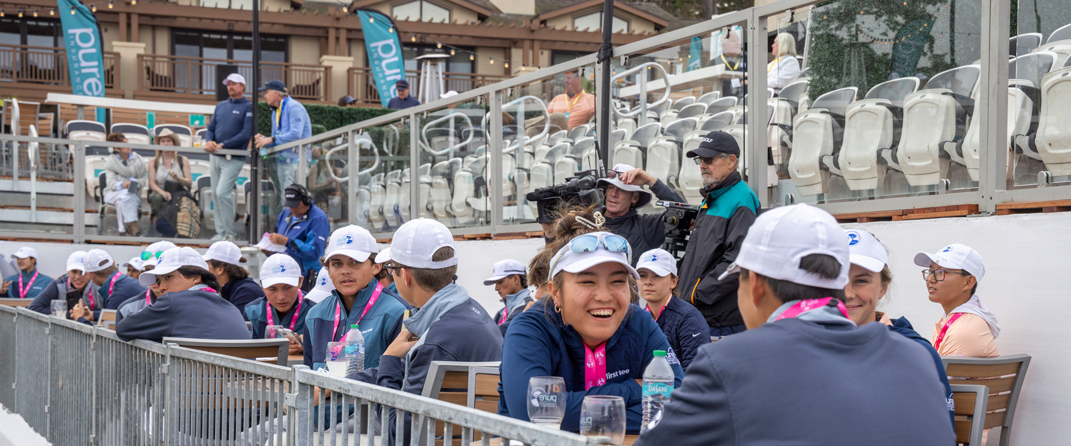 First Tee participants wearing white caps and pink lanyards laughing together in a stadium seating area