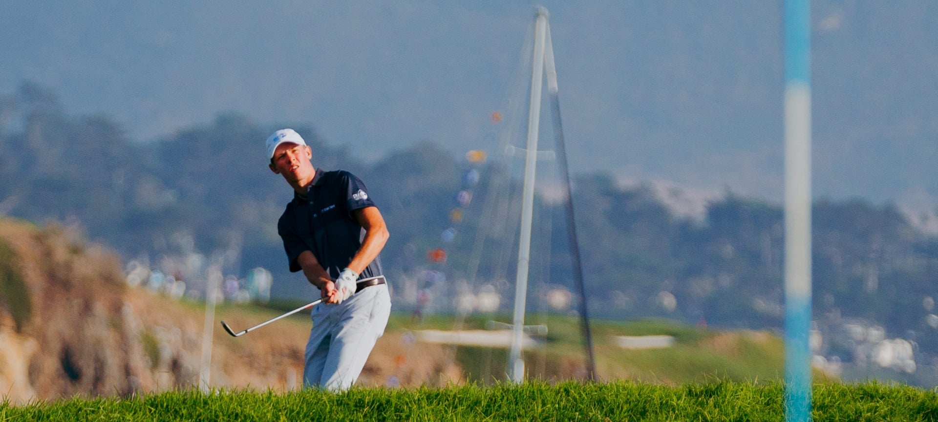 A young male golfer in a navy polo watches his shot fly toward a coastal backdrop at Pebble Beach