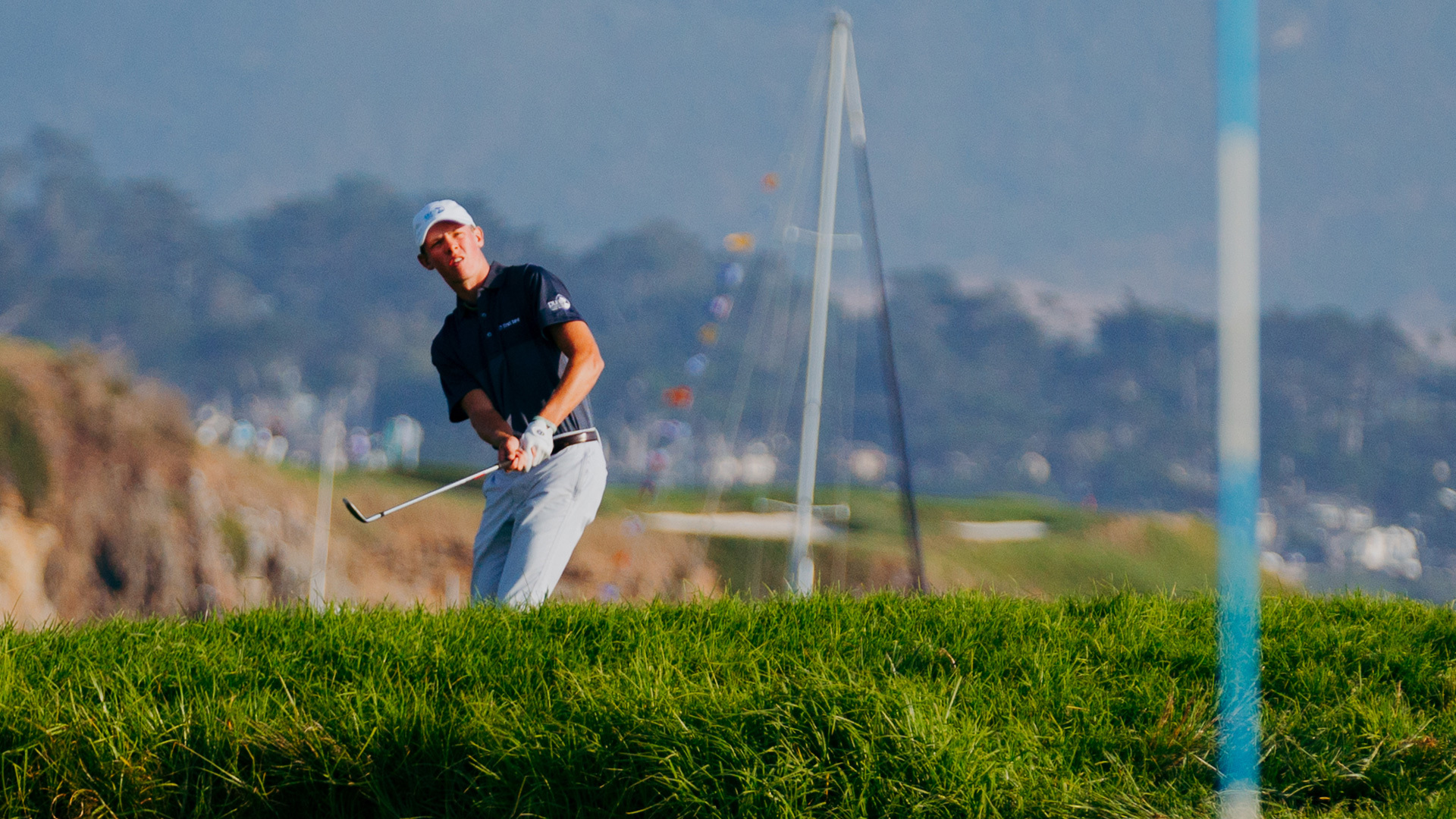 A young male golfer in a navy polo watches his shot fly toward a coastal backdrop at Pebble Beach